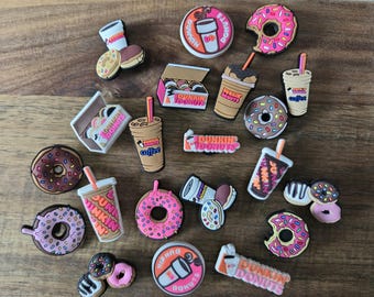 May include: A selection of colourful shoe charms featuring donuts, coffee cups, and the Dunkin' Donuts logo. The charms include pink iced donuts, iced coffee, and boxes of donuts. The charms are on a wooden surface.