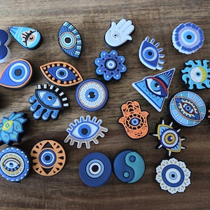 Puede incluir: Colección de dijes para zapatos azules, blancos y naranjas con varios diseños, incluyendo ojos, manos y patrones florales. Los dijes están dispuestos sobre una superficie de madera.
