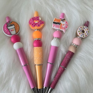 May include: Four decorative pens with unique designs. One pen features a Dunkin' Donuts logo, another a donut, and a third reads "She Wants The." The fourth pen has a donut design. Each pen has a pink barrel, silver accents, and a decorative top.