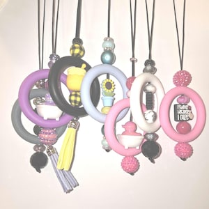May include: A collection of colorful silicone necklaces with various charms, including a sunflower, a bathtub, a duck, and a black square with the text "I swear because I care".