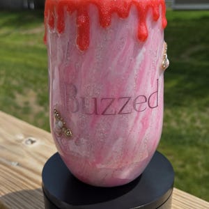 May include: A pink and white tumbler with the word "Buzzed" printed on the side. The top of the tumbler is decorated with red glitter drips, a honey dipper, and decorative jewels. The tumbler sits on a black base.