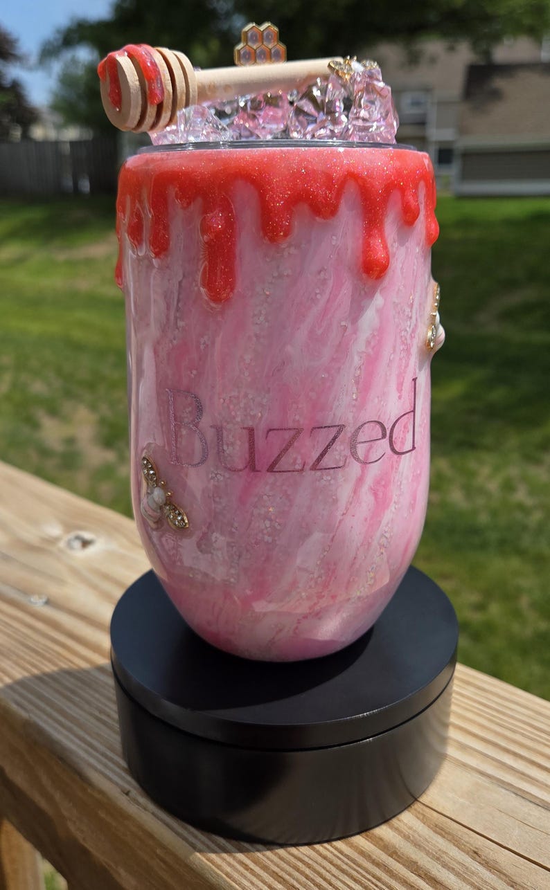 May include: A pink and white tumbler with the word "Buzzed" printed on the side. The top of the tumbler is decorated with red glitter drips, a honey dipper, and decorative jewels. The tumbler sits on a black base.