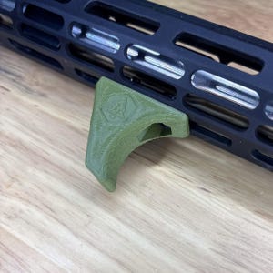 May include: An olive green angled foregrip with a textured, 3D-printed finish and a small hexagonal logo embossed on the side. The accessory is securely attached to the underside of a black, ventilated metal handguard featuring elongated slots.