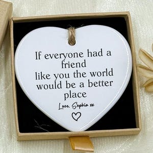 May include: A heart-shaped white ceramic ornament with a printed message inside a small gift box. The box is tan with the words "Hand made" on the lid. The ornament reads "If everyone had a friend like you the world would be a better place."