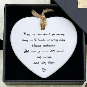 May include: A white heart-shaped ornament with a jute string, displayed in a black gift box. The ornament has a poem printed on it. The box is open, revealing the ornament, and is decorated with a "Hand made" label and blue flowers.