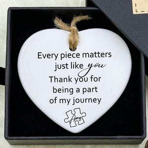 May include: A white heart-shaped ornament with a jute string, inside a black gift box. The ornament has the text "Every piece matters just like you. Thank you for being a part of my journey." The box is labeled "Hand made."