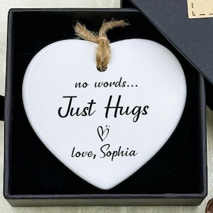 Puede incluir: Un adorno blanco en forma de corazón con las palabras "Just Hugs" y "love, Sophia" en una caja de regalo negra. La caja tiene una etiqueta "Hand made" y detalles de flores azules. El adorno está atado con hilo.
