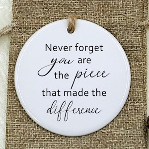 May include: A white, circular ceramic ornament inscribed with the words "Never forget you are the piece that made the difference" in black script. The ornament is presented in a small hessian bag with a drawstring, with decorative elements.