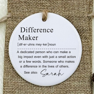 May include: A white, round ornament with the text "Difference Maker" and a definition. The ornament is hanging from a jute bag. The definition includes the name "Sarah". The background includes flowers and rings.