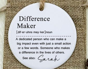 May include: A white, circular ornament with the words "Difference Maker" and a definition printed on it. The ornament is displayed on a jute bag. The definition includes the name "Sarah". The background includes flowers and rings.