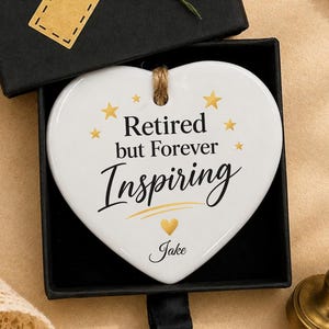 May include: A white heart-shaped ceramic ornament inside a black gift box. The ornament features the text "Retired but Forever Inspiring" with gold stars, a gold heart, and the name "Jake" in black script. The box lid has two blue flowers.
