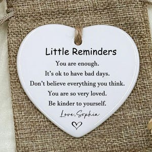 May include: A white heart-shaped ceramic ornament with the text "Little Reminders" and inspirational quotes, hanging on a jute string. It rests on a burlap bag, next to a small drawstring bag. The ornament is a thoughtful gift.