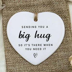 Puede incluir: Un adorno blanco en forma de corazón con el texto "Sending you a big hug" en letras negras. El adorno está sobre una bolsa de arpillera, con una bolsa con cordón a juego a la izquierda. El fondo incluye detalles florales.