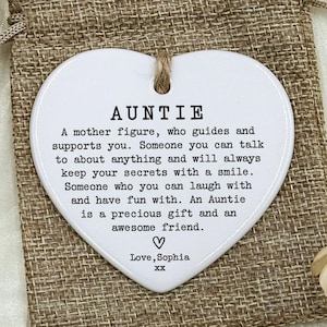 May include: A white heart-shaped ceramic ornament with the word "AUNTIE" and a message about aunties. The ornament hangs from a jute string and rests on a burlap bag. A small burlap gift bag is also in the image.