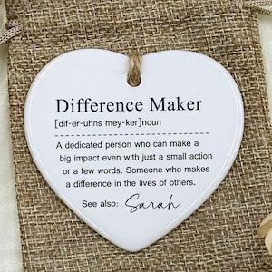 Puede incluir: Un adorno de cerámica blanco en forma de corazón con el texto "Difference Maker" y una definición. El adorno cuelga de una cuerda de yute y descansa sobre una bolsa de arpillera. También se ve una bolsa de arpillera más pequeña.