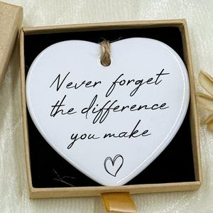 May include: A heart-shaped ceramic ornament with the text "Never forget the difference you make" inside a small gift box. The box is tan with a "Hand made" label and a small floral accent. A decorative gift item.