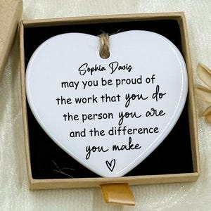 May include: A heart-shaped white ceramic ornament with a printed message, presented in a small, open gift box. The box is tan with a "Hand made" label. The ornament has a jute string for hanging.
