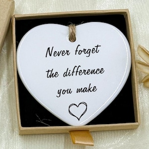 May include: A heart-shaped white ceramic ornament with the text "Never forget the difference you make" inside a small gift box. The box is tan with a decorative flower and the words "Hand made".