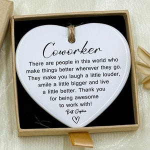 May include: A heart-shaped white ceramic ornament with the word "Coworker" and a message inside a small gift box. The box is tan with a "Hand made" label and a small floral accent.