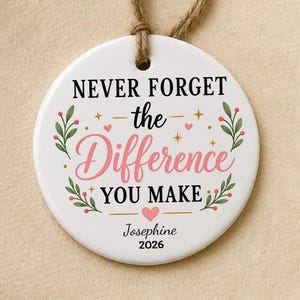 May include: A round white ceramic ornament with a jute twine loop. The text reads NEVER FORGET the Difference YOU MAKE, Josephine 2026. It features green leafy sprigs, small pink hearts, and gold stars printed on the front surface of the ornament.