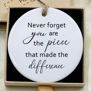 May include: A white ceramic ornament with the text "Never forget you are the piece that made the difference" in black script. The ornament is in a gift box with a "Hand made" tag. Decorative flowers are placed around the box.