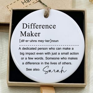 Pode incluir: Um ornamento de cerâmica branco e redondo com o texto "Difference Maker" em preto, apresentado numa pequena caixa de presente. O ornamento inclui uma definição e o nome "Sarah". A caixa é castanha com uma etiqueta "Hand made".
