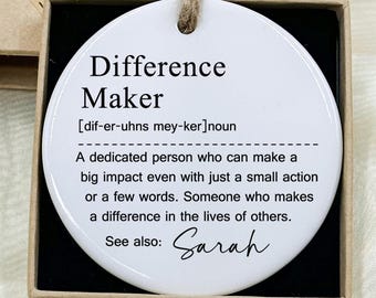 May include: A white, circular ceramic ornament with the text "Difference Maker" in black, presented in a small gift box. The ornament includes a definition and the name "Sarah". The box is brown with a "Hand made" label.