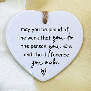 Könnte beinhalten: Weißes herzförmiges Keramikornament mit einer Juteschnur zum Aufhängen. Das Ornament trägt eine Botschaft in schwarzer Schrift: "may you be proud of the work that you do, the person you are and the difference you make" mit einem Herzsymbol.