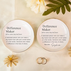 May include: Two white ceramic ring dishes with gold rims, each featuring the text "Difference Maker" and a definition. One dish includes the name "Sophia." Decorative flowers and rings are also present.