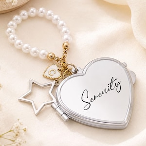 Può includere: Uno specchietto da borsa argentato a forma di cuore con la scritta Serenity in corsivo nero. Lo specchio è fissato a un cinturino di perle bianche con una catenella dorata, un piccolo ciondolo a cuore con la lettera C e una stella argentata.