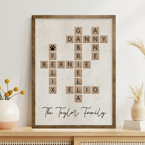May include: A framed family name art piece. The artwork features a crossword puzzle design with names spelled out using Scrabble tiles. The names include "Danny", "Bernie", and "Elio". The text "The Taylor Family" is written below the puzzle.