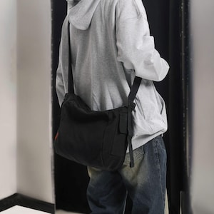 May include: A black canvas messenger bag with a long adjustable strap worn over the shoulder. The bag has a flap closure and a small pocket on the front.