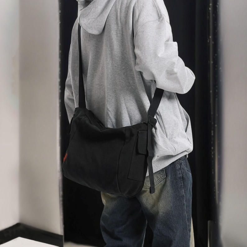 May include: A black canvas messenger bag with a long adjustable strap worn over the shoulder. The bag has a flap closure and a small pocket on the front.