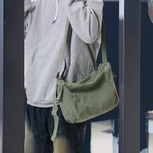 May include: A person wearing a gray hoodie and dark blue jeans is carrying a green canvas shoulder bag with a long strap.