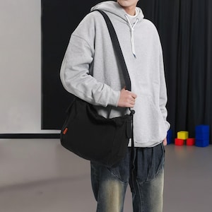 May include: A person wearing a gray hoodie with a black canvas shoulder bag. The bag has a long strap and is worn over the person's shoulder.