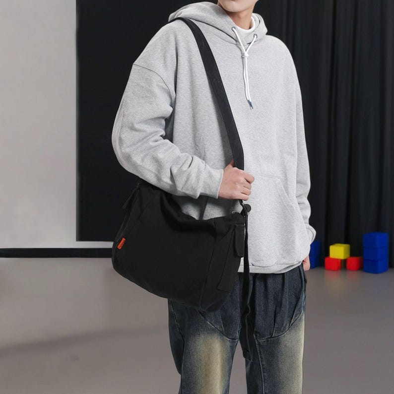 May include: A person wearing a gray hoodie with a black canvas shoulder bag. The bag has a long strap and is worn over the person's shoulder.