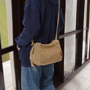 May include: A tan canvas shoulder bag with a zipper closure and a long strap. The bag is worn over the shoulder of a person wearing a blue hoodie and jeans.