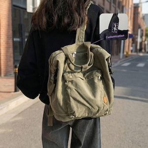 May include: An olive green canvas backpack with multiple pockets and a top handle. The bag has a rectangular shape with a flap and buckle closure. A small tag with the word "april" is visible on the strap. The backpack is worn over the shoulder.
