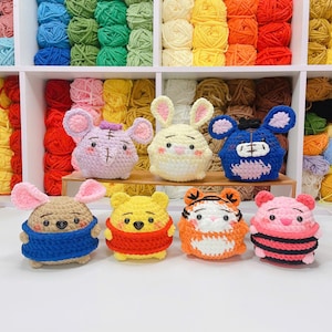 May include: A collection of eight crocheted stuffed toys, including a brown bear, a pink pig, a yellow bunny, a blue bear, a brown bear wearing a blue scarf, a yellow bear wearing a red sweater, a tiger wearing a white and orange striped sweater, and a purple elephant.