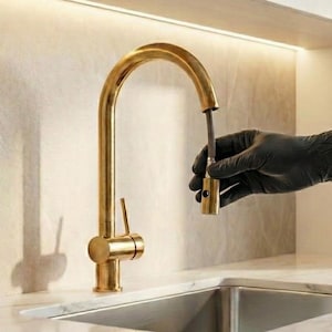 Pode incluir: Uma torneira de cozinha dourada com um bico de arco alto. A torneira está instalada em uma bancada branca com uma pia de aço inoxidável. Uma pessoa com uma luva preta está segurando a cabeça da torneira.