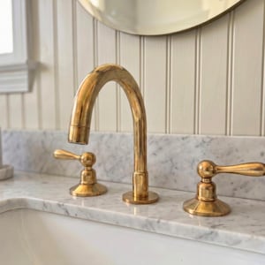 May include: A gold-toned bathroom faucet with a curved spout and two lever handles. The faucet is mounted on a white marble countertop. A brown glass bottle with a black pump is to the right.