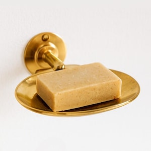 May include: A brass-coloured soap dish with a wall mount, holding a rectangular bar of soap. The soap dish has a simple, elegant design, suitable for a bathroom or kitchen. The soap bar is light brown.