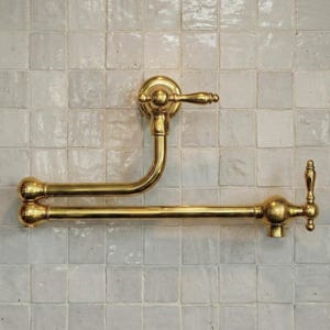 Könnte beinhalten: Ein goldfarbener Topffüller-Wasserhahn mit gebogenem Auslauf und dekorativen Griffen. Der Wasserhahn ist an einer weiß gefliesten Wand montiert und zeigt ein klassisches Design. Der Wasserhahn ist aus Messing.