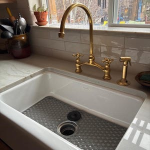 Unlacquered brass bridge kitchen faucet with cross handles and side sprayer over white farmhouse apron sink with marble countertop.