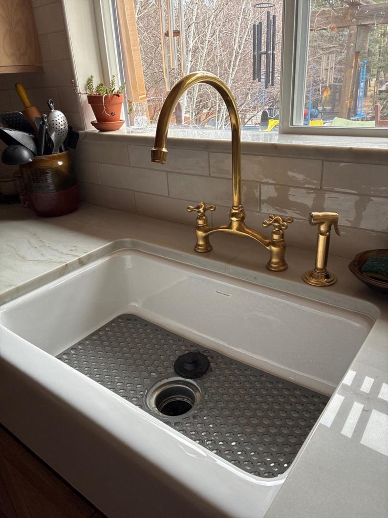 Unlacquered brass bridge kitchen faucet with cross handles and side sprayer over white farmhouse apron sink with marble countertop.