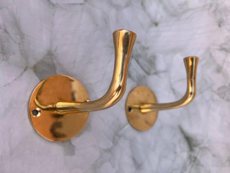 May include: Two brass-colored wall hooks with a curved design. Each hook is mounted on a circular base. The hooks are a shiny gold color and are set against a white and gray marbled background.