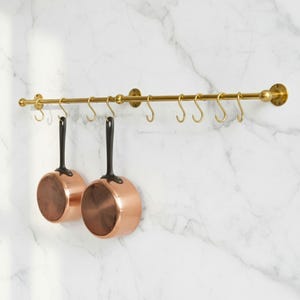 May include: A brass pot rack with eight hooks, holding two copper-bottomed saucepans with black handles. The rack is mounted on a white marble-patterned wall, offering a stylish kitchen storage solution.