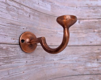 May include: A copper-colored wall hook with a curved design. The hook has a circular base with a single screw hole and a curved arm that leads to a cup-shaped end. The hook is mounted on a wooden surface.