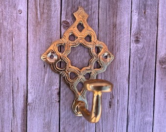 May include: Ornate brass wall hook with a decorative diamond-shaped backplate and a curved hook. The backplate features an openwork design with intricate patterns. The hook is mounted on a wooden surface.