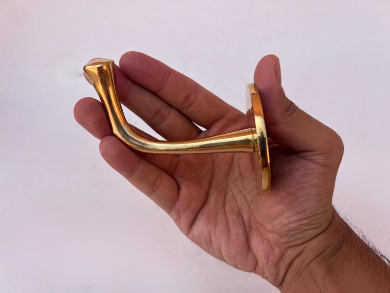 Hand holding brass wall hook against white background.